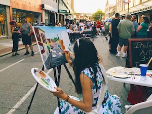 Woman painting on a canvas in the middle of a busy street.